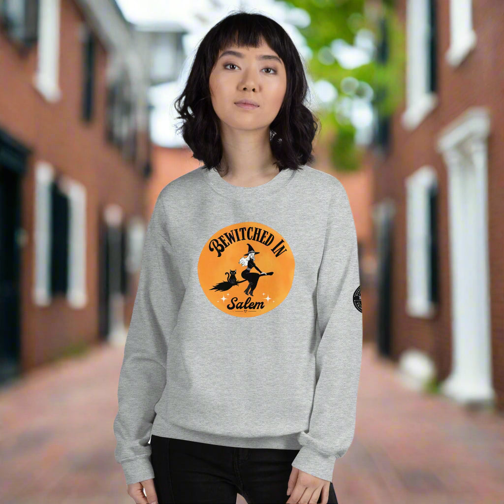 woman wearing a ight crew neck sweatshirt that has an orange moon, witch and black cat on a broom, it says "Bewitched in Salem" on it.