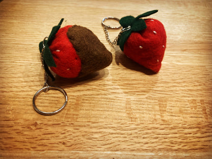 Two strawberry-shaped keychains on a wooden surface one is chocolate dipped made of felt